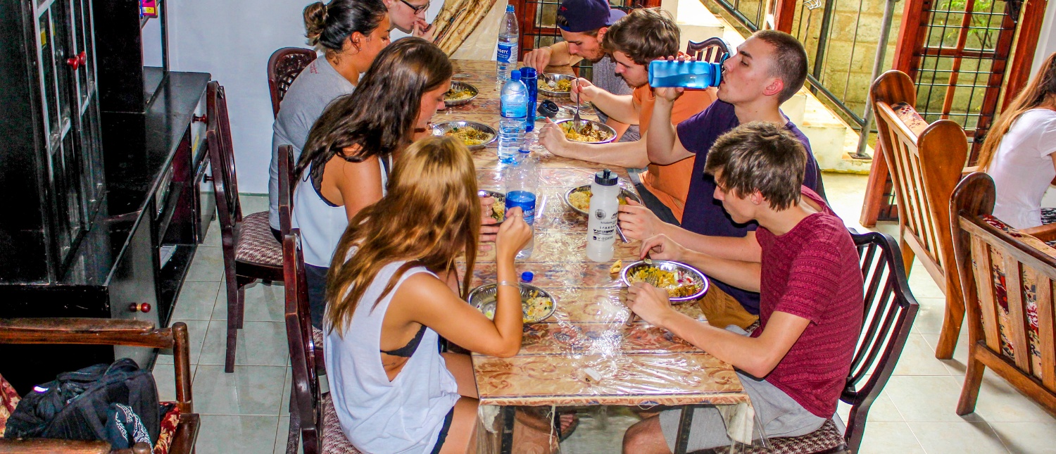 A group of young travelers share a casual meal around a long table indoors.